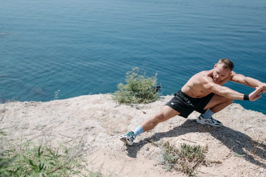 Shirtless man doing a stretch on a cliff overlooking the ocean in daylight.